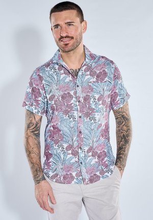 Mann mit Tätowierungen, trägt ein kurzärmeliges Hemd mit lila-blauem Blumenmuster und helle Hosen, steht mit den Händen in den Taschen.