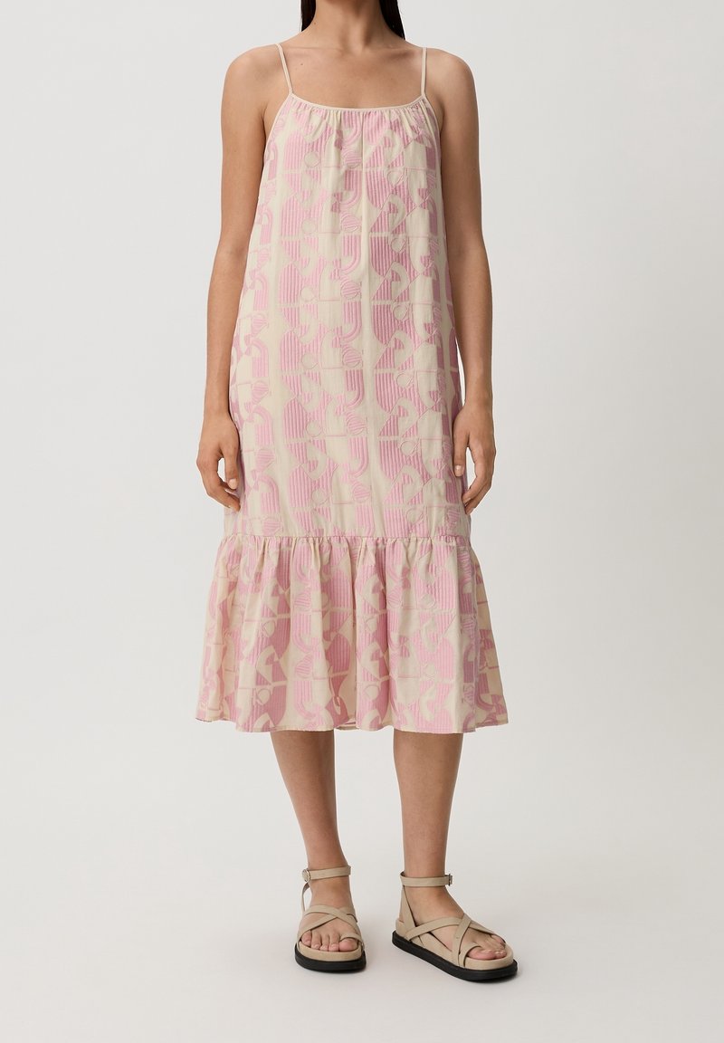 Femme portant une robe sans manches, longueur genoux, rose et beige à motifs avec un ourlet à volants et des sandales plates beiges avec brides à la cheville.
