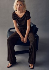 Tmavě hnědý třpytivý overal s lěmovým výstřihem, krátkými rukávy a vázáním v pase. Kombinováno s černými sandály na pásech na modré židli.