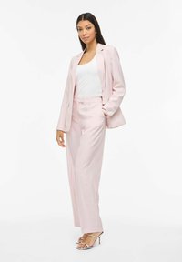 Costume tailleur rose avec un blazer à simple boutonnière et un pantalon ample. Un haut blanc en dessous et des talons argentés à brides complètent le look.