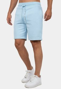 Shorts en mélange de coton bleu clair avec une taille à cordon, poches latérales et un logo brodé discret en bas à gauche. Texture lisse.