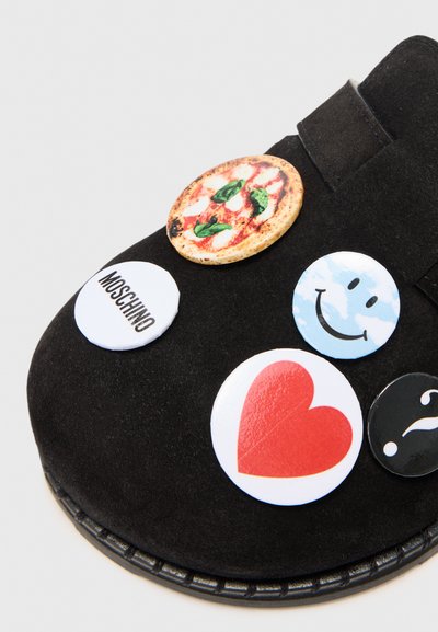 Zapato de ante negro con varios parches circulares: una porción de pizza, una cara sonriente, un corazón, un signo de interrogación y el texto "Moschino".