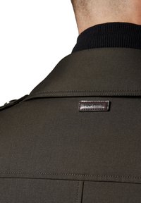 Ciemnozielony płaszcz z teksturowanej tkaniny, z czarnym prążkowanym kołnierzem i markową brązową skórzaną metką z tyłu.