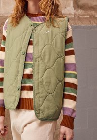 Person i grön quiltad Nike-väst över randig stickad tröja med färgerna lila, grön, kräm, brun och gul, tillsammans med ljusa beige byxor.