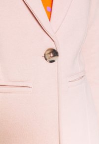 Ljusrosa blazer med en dekorativ knapp, två framfickor och ett texturerat tyg med subtila sömnadsdetaljer.