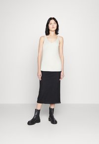 Weißes, geripptes Tanktop, schwarzer Sliprock mit Spitze, und schwarze Ankle Boots. Das Model steht vor einem hellgrauen Hintergrund.