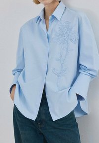 Cămașă albastru deschis cu nasturi și mâneci lungi, cu un detaliu brodat floral pe partea stângă. Confecționată dintr-un material fin.