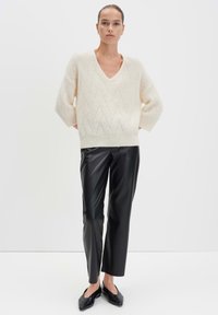 Frau steht mit den Händen hinter dem Rücken, trägt einen weißen Strick-V-Ausschnitt-Pullover, schwarze gerade Lederhosen und schwarze spitze Ballerinas.