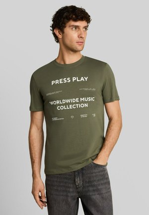 Mężczyzna o kręconych ciemnych włosach, ubrany w oliwkowo-zielony t-shirt z białym napisem ""Press Play Worldwide Music Collection,"" i ciemnoszare jeansy.