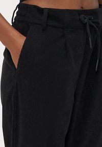 Black corduroy pants with a drawstring waist, side pockets, and a ridged texture. Features detailing on the waistband and pockets.