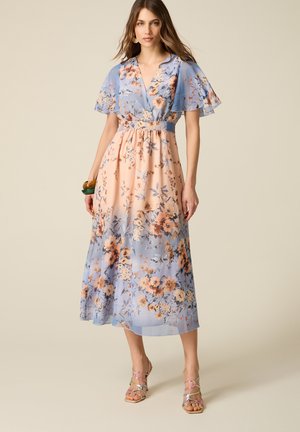 Frau trägt ein Midikleid mit Blumenmuster, flatternden Ärmeln und Pastellblau- sowie Pfirsichtönen, kombiniert mit Riemchenabsätzen und Armbändern.