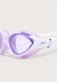 Προστατευτικά γυαλιά κολύμβησης με μοβ απόχρωση, λευκά ρυθμιζόμενα λουράκια και μικρό μοβ λογότυπο στο πλάι, τοποθετημένα σε λευκή επιφάνεια.