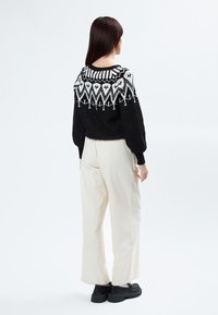 Schwarzer Strickpullover mit weißen geometrischen Mustern an der Passe, kombiniert mit hochgeschnittenen cremefarbenen Hosen und klobigen schwarzen Schuhen.