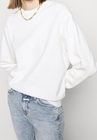 Personne portant un pull long à manches longues en côte blanche, un jean taille haute bleu clair, et un collier en chaîne dorée sur un fond uni.