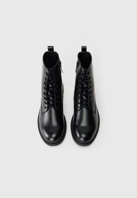 Botines de piel negra con textura suave, puntera redonda, cordones en la parte delantera y cremallera lateral para facilitar el uso. Incluyen una pestaña trasera para ayudar a calzarlos.