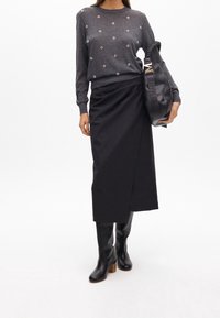 Femme portant un pull gris avec de petits détails floraux, une jupe midi asymétrique noire, des bottes noires montant jusqu'aux genoux, et tenant un sac à main noir.
