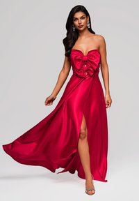 Abito da sera in satin rosso con design senza spalline, grande dettaglio a forma di fiocco sul davanti e spacco alto. Texture liscia con gonna fluente.