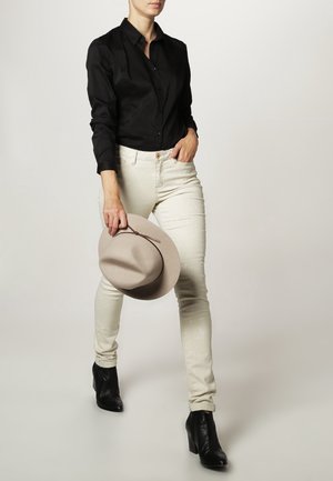 Black button-up shirt, cream skinny jeans, and black ankle boots. Holding a light beige hat. Simple, fitted design with clean lines.