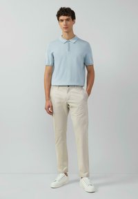 Hellblaues Polohemd mit kurzen Ärmeln und drei Knöpfen, kombiniert mit beigen Hosen und weißen Sneakers. Glatte Textur und figurbetontes Design.