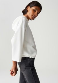 Vrouw met kort, donker haar die een gestructureerde witte hoodie en donkergrijze jeans draagt, staande zijwaarts tegen een effen lichte achtergrond.