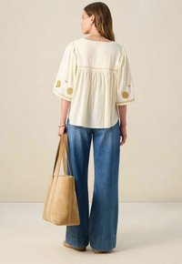 Cremfarbene Bluse mit gestickten goldenen Akzenten, ausgestellten Ärmeln, kombiniert mit hochgeschnittenen blauen Jeans und einer hellbraunen Wildleder-Shopper-Tasche.