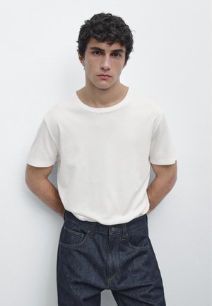 Jonge man met krullend donker haar, gekleed in een eenvoudig wit T-shirt dat in een donkerblauwe jeans is gestopt, staand tegen een witte muur.