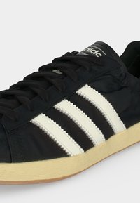 Μαύρο αθλητικό παπούτσι Adidas με λευκές τρεις ρίγες, μαύρα κορδόνια και μπεζ σόλα, στραμμένο για να δείξει πλευρικές και επάνω λεπτομέρειες.
