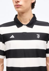 Άτομο που φοράει μπλούζα πόλο με μαύρες και λευκές ρίγες, με τα λογότυπα της Adidas και της Juventus στο στήθος, όρθιο μπροστά σε λευκό φόντο.