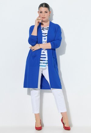 Vrouw staand tegen een witte muur, dragend een blauwe open cardigan, een shirt met patroon, witte broek en rode hoge hakken, met een hand bij haar gezicht.