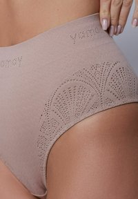 Högmidjad beiga underkläder med ett strukturerat mönster och dekorativa laserskurna detaljer längs benkanten. Logotypen är synlig på midjebandet.