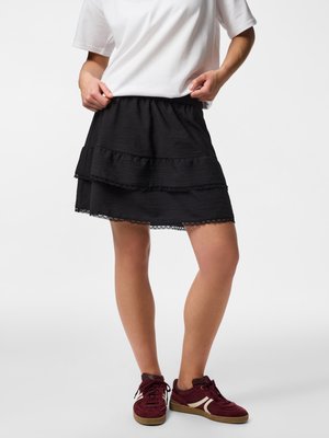Personne portant un t-shirt blanc, une jupe noire à volants avec une bordure en dentelle et des baskets bordeaux, debout devant un fond clair et uni.