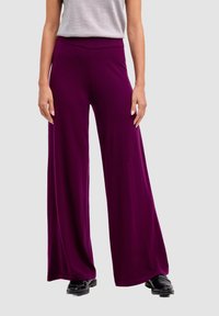 Donna che indossa pantaloni viola a vita alta e gamba larga, con scarpe nere e un top senza maniche grigio chiaro, su uno sfondo neutro.