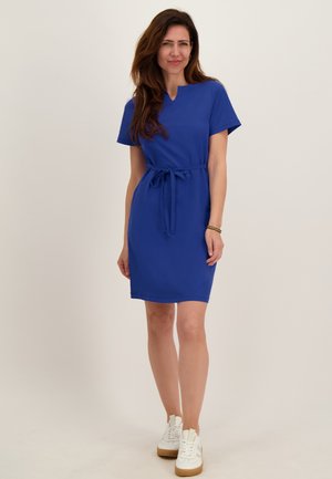 Vrouw draagt een koningsblauwe jurk tot op de knie met korte mouwen en een tailleceintuur, gecombineerd met witte sneakers, staand tegen een effen achtergrond.