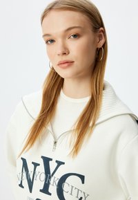 Sudadera blanca con cremallera y texto en navy "NYC" y "NEW YORK CITY" en diferentes tamaños. Cuenta con un cuello de canalé y una textura suave.