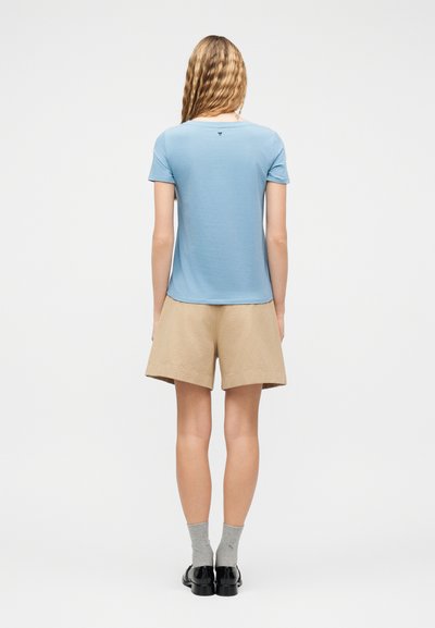 Γυναίκα με κυματιστά ξανθά μαλλιά που φοράει ανοιχτό μπλε κοντομάνικο πουκάμισο, μπεζ σορτς, γκρι κάλτσες και μαύρα παπούτσια, στέκεται με την πλάτη προς τα εμάς.