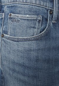 Blå denimjeans med en blekt textur, flera fickor, nitar och söm detaljer. Materialet är mjukt och något slitet.
