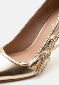 Κοντινή λήψη ενός μεταλλικού χρυσού μπαλαρίνου με στρογγυλεμένη μύτη και διακοσμητικό φιόγκο στο πλάι.