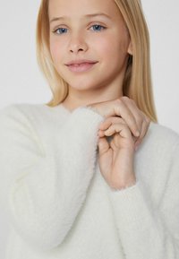 Flauschiger weißer Pullover mit einer weichen Textur und gerippten Bündchen, der einen entspannten Schnitt bietet. Die Hände sind sanft am Halsbereich zusammengelegt.