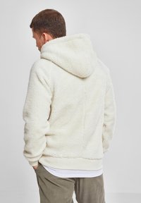 Cremefarget fleece-hettegenser med teksturert ytre, over en hvit skjorte, parret med olivengrønne bukser. Har en stor hette og elastiske mansjetter.