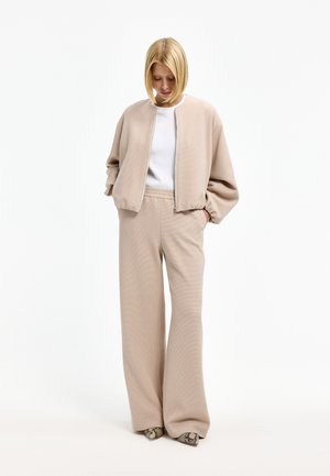 Person wearing beige oversized zip-up jacket and matching wide-leg pants over white top, standing with hands in pockets, looking down.