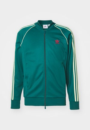 Žalias sportinis švarkas su užtrauktuku, trys geltonos juostelės ant rankovių ir raudonas logotipas ant krūtinės. Sklandus audinio paviršius.