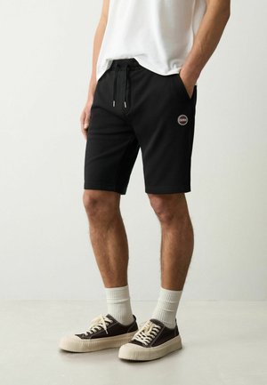 Shorts neri realizzati in un tessuto morbido con cintura elasticizzata, tasche laterali e una toppa con logo rotondo. Indossati con calzini bianchi e scarpe scure.