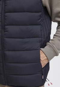 Mornarica puffer prsluk s kvadratnim uzorkom, zatvaranje na patentni zatvarač i bočna džep. Elastične manžete i naglasci sđu na crvenoj i bijeloj detaljima.