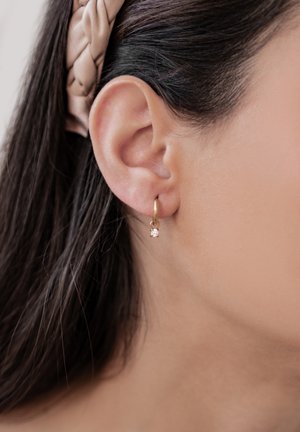 Goldene Creolen mit einem kleinen, klaren Edelsteinakzent, präsentiert am Ohr eines Modells mit dunklem Haar und einem neutralen Hintergrund.