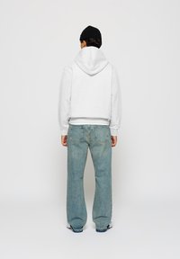 Mann iført en svart lue, lys grå hettegenser, løse blå jeans og hvite joggesko, vendt bort fra kameraet mot en enkel lys bakgrunn.