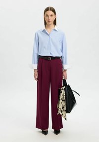Hellblaue gestreifte Button-Up-Bluse, hochtaillierte kastanienbraune weite Hosen, schwarzer Gürtel, schwarze Handtasche mit einem gepunkteten Schal-Akzent, spitze Schuhe.
