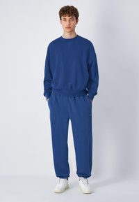 Blauwe sweatshirt en bijpassende joggingbroek van zachte stof. De outfit heeft een casual pasvorm met geribbelde boorden en een tailleband. Witte sneakers.