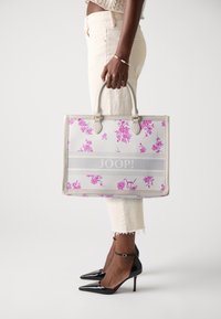 Borsa tote bianca con stampa floreale rosa e striscia grigia. Design strutturato, doppio manico superiore e un logo. I pantaloni sono chiari, le scarpe sono tacchi neri.