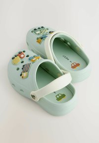 Svetlozelené gumové dreváky majú biely remienok, vetraný dizajn a hravé 3D ozdoby v podobe hračiek a áut na vonkajšej strane.