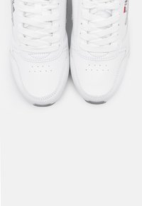 Chaussures de sport blanches avec une tige en cuir lisse, des panneaux en mesh et des lacets ronds. Elles comportent des perforations pour la ventilation et des semelles en caoutchouc.